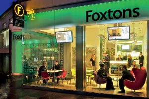 Foxtons estate agency branch with modern, brightly lit interior. Staff and customers are seated at tables and chairs, engaging in discussions. The exterior features large glass windows, illuminated signage, and digital displays showing property listings