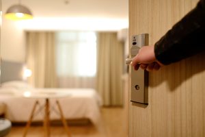 Image depicts someone opening a hotel room door to reveal a warm, inviting room, cream colour curtains, bed and wooden floor.