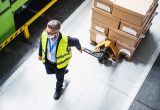 A worker wearing a reflective safety vest and face mask pulls a pallet jack loaded with stacked cardboard boxes through a warehouse.