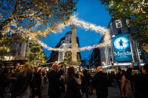 A busy evening scene in London’s West End decorated with festive lights. Crowds of people walk and gather around Seven Dials’ central column, with trees and buildings illuminated by sparkling white lights. A large “Matilda the Musical” theatre billboard is visible on the right-hand side