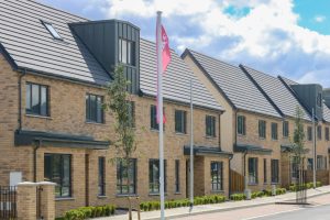 A row of modern, newly built townhouses with brick facades and dark grey tiled roofs. Each unit has large windows and small front gardens with young trees. A pink flag on a pole stands in front. The sky is partly cloudy with blue patches.