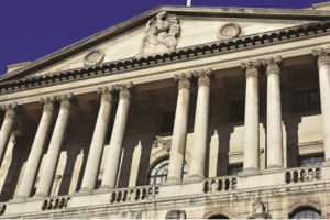 A view of the historic Bank of England building in London, featuring grand neoclassical architecture with tall Corinthian columns, arched windows, and detailed stone carvings under a blue sky.
