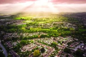 An aerial view of a suburban neighbourhood surrounded by green trees and open countryside. Sunrays break through the e clouds, casting a glowing light over the landscape. The neatly arranged houses and winding roads contrast with the natural greenery and farmland in the distance.