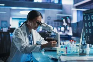There is a scientist in a modern looking science lab. She is wearing a lab coat and looking into a microscope.