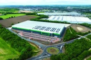 Tritax asset. Aerial view of a large modern logistics warehouse with a white roof partially covered in solar panels. The building features green and blue accents and is surrounded by landscaped areas, parking spaces, and access roads. Nearby are other industrial buildings, fields, and a roundabout leading to the facility, all set in a semi-rural environment