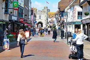 A busy pedestrian street in a historic town, lined with small shops and restaurants. Colourful bunting hangs overhead, and people walk along a brick-paved road. In the distance, a medieval stone gate tower with a flag on top stands at the end of the street. On the right, a woman pushes a pram, while others stroll, shop, and talk in the sunny daytime scene.
