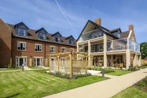 **Alt text:** Modern brick residential building with multiple stories and dormer windows, featuring balconies with glass railings, a landscaped lawn, and a wooden pergola in the foreground under a clear blue sky.