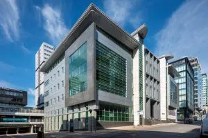 A modern multi‑storey office building on a city street corner, featuring large green‑tinted glass windows and light grey cladding. The building has sharp angular lines and a prominent overhanging roof. Surrounding it are other contemporary high‑rise buildings under a bright blue sky with wispy clouds.