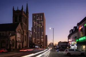 Lord Street - picture shows twilight city street scene showing a tall modern residential tower next to an older stone church with two spires. Light trails from passing vehicles run along the road in the foreground. On the right side of the street are small shops with bright neon signs. The sky is a gradient of purple and orange as the sun sets behind the skyline in the distance.
