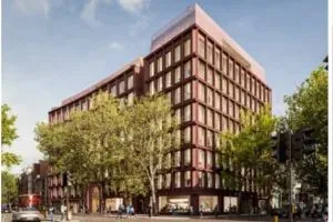 CGI of Xylo shows a large, modern building with a reddish-brown exterior and many tall windows sits on a street corner. Mature trees with green foliage line the front of the building, partially obscuring some of the façade. People are walking along the pavement and a few cars are visible on the road. The sky is bright with scattered clouds