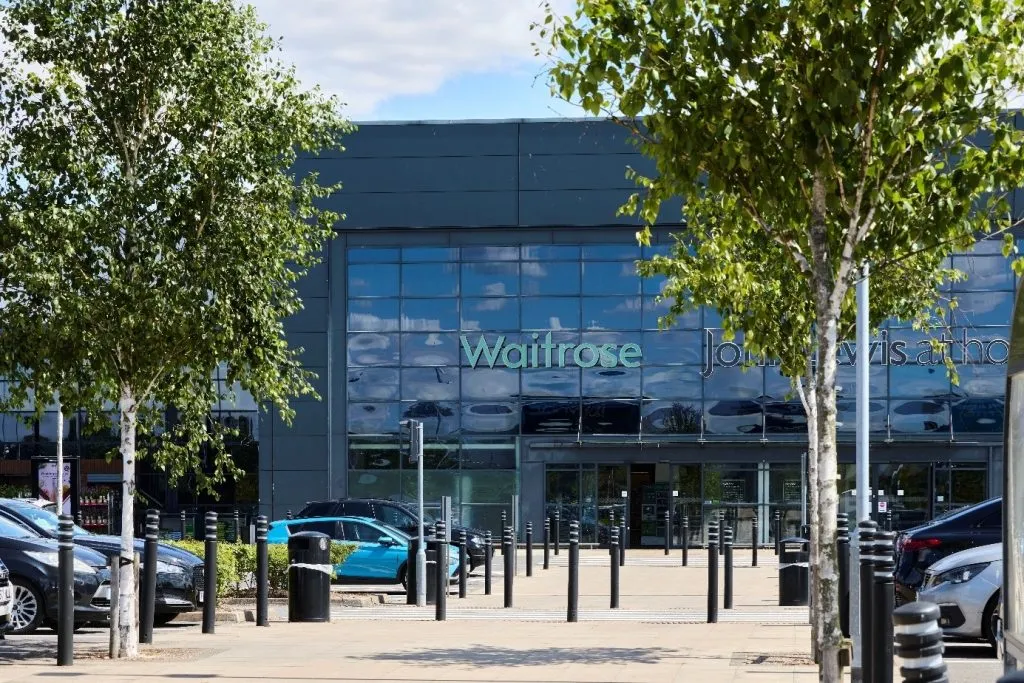 Exterior of a Waitrose supermarket with large glass windows, viewed from a car park lined with trees and parked vehicles.
