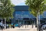Exterior of a Waitrose supermarket with large glass windows, viewed from a car park lined with trees and parked vehicles.