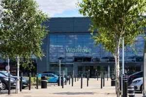 Exterior of a Waitrose supermarket with large glass windows, viewed from a car park lined with trees and parked vehicles.